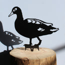 The Laysan Duck by Metalbird Canada - CC is a black metal duck silhouette mounted on a wooden log, casting a striking shadow on light blue—ideal as unique garden art or a thoughtful gift.