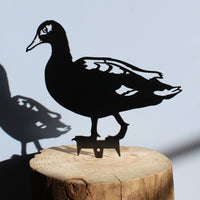 The Laysan Duck by Metalbird Canada - CC is a black metal duck silhouette mounted on a wooden log, casting a striking shadow on light blue—ideal as unique garden art or a thoughtful gift.