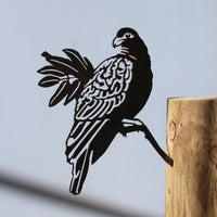 The Manumea by Metalbird Canada - CC is a corten steel parrot silhouette perched on a wooden post, casting its shadow against a plain light blue background.