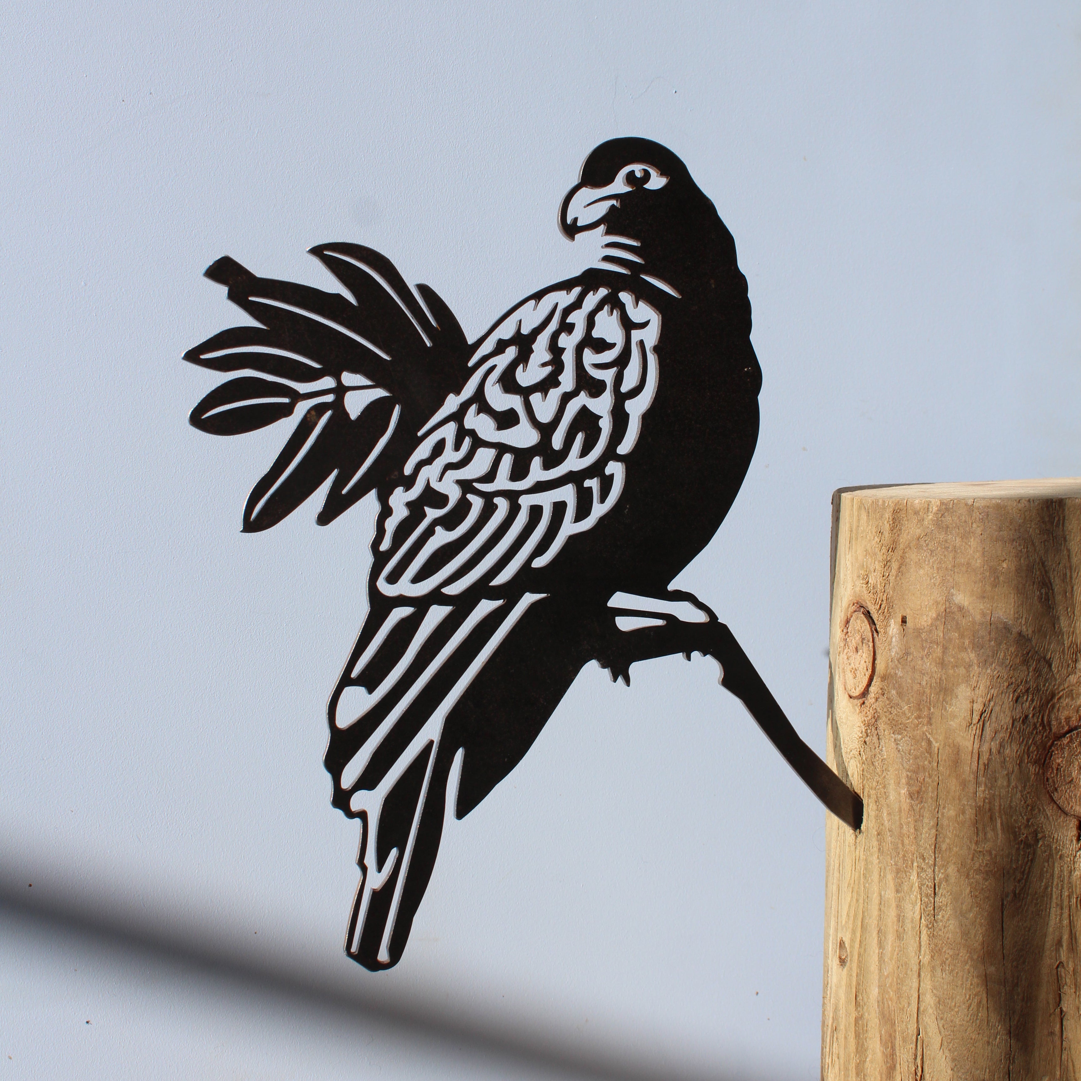 The Manumea by Metalbird Canada - CC is a corten steel parrot silhouette perched on a wooden post, casting its shadow against a plain light blue background.