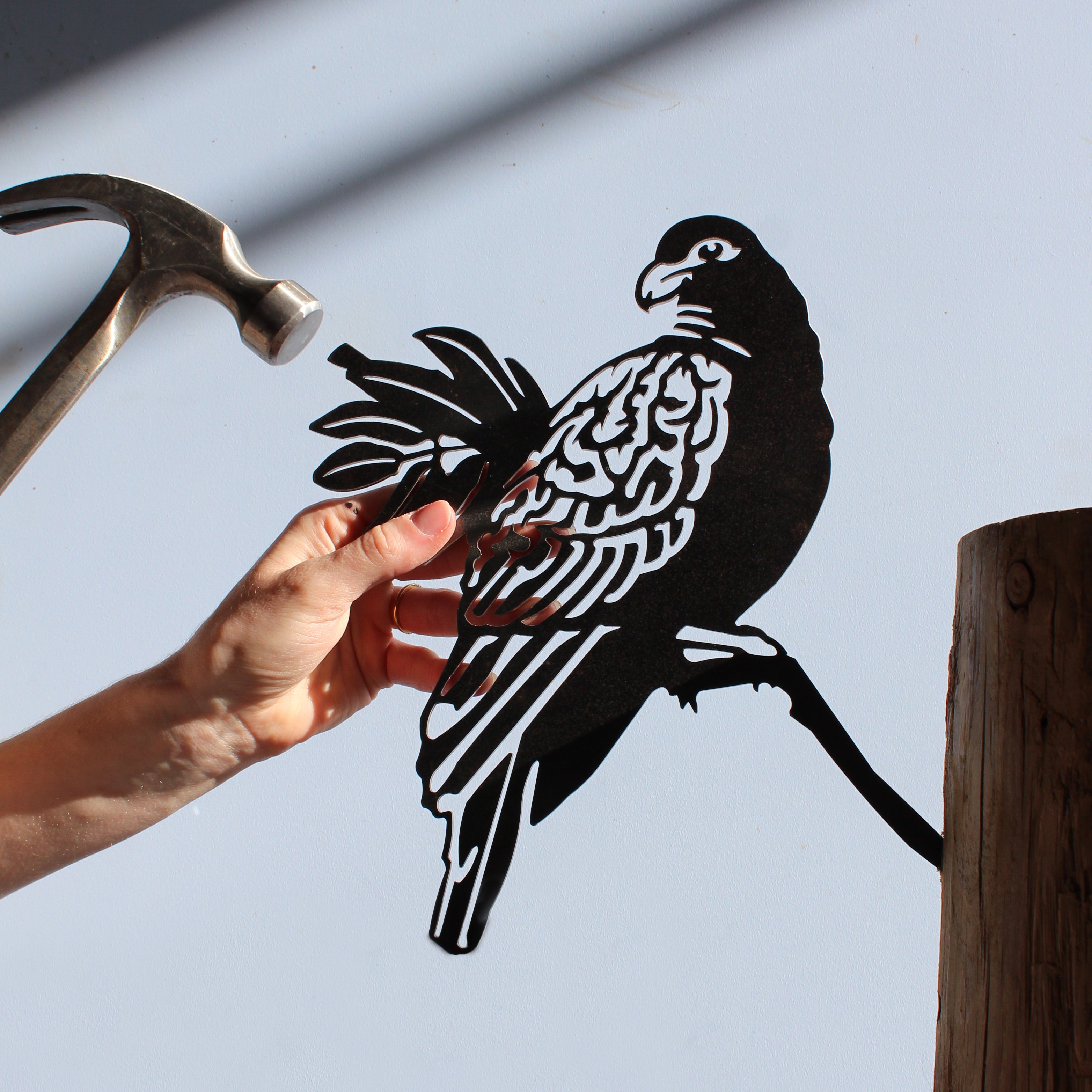 A hand holds the Manumea, a detailed black bird silhouette by Metalbird Canada - CC, perched on a branch against a light background. A hammer and wooden post nearby inspire striking, unique garden art.