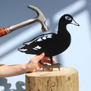 A hand holds the Metalbird Canada - CC Laysan Duck, a black metal garden art silhouette, against a wood stump as another hand raises a hammer. The light blue background features striking shadows.