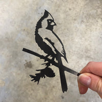 A hand holds the Metalbird Canada "Cardinal Mom & Mini Set," a thin black metal cutout of cardinal birds on a branch—an ideal gift for bird lovers. The background is a blurred gray surface.