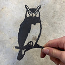 A hand holds Metalbird Canada's Mini Owl, a flat black metal silhouette perched on a branch—ideal as garden art—set against a light gray, textured background.