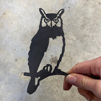 A hand holds Metalbird Canada's Mini Owl, a flat black metal silhouette perched on a branch—ideal as garden art—set against a light gray, textured background.