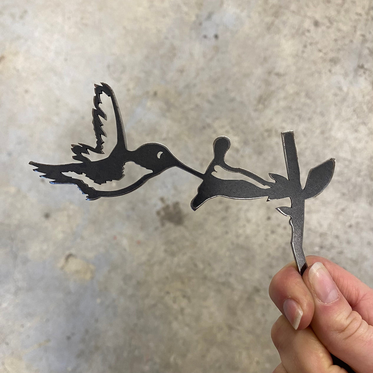 Hand holding the Mini Hummingbird steel silhouette beside its flower against a textured grey wall.