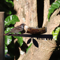 A sparrow perches on the Metalbird Canada Stick Bird Feeder with a banana slice, set against green leaves and tree bark. The partial brand name appears on this unique corten steel garden art piece.