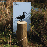 A Laysan Duck silhouette by Metalbird Canada - CC, crafted from metal, is mounted on a wooden log near tall grasses and reeds by a pond, in front of a BirdLife International sign.