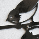 A close-up of Metalbird Seconds by Metalbird Canada - Seconds, a black corten steel wall art shaped like a bird on a branch, mounted on a textured white wall—an ideal unique gift.