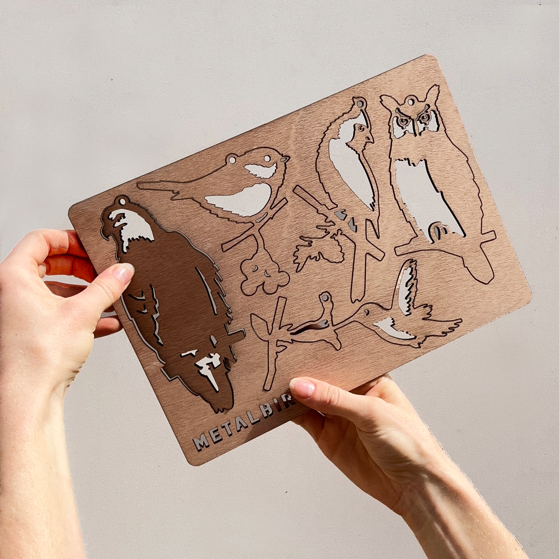 A person holds a Wooden Hanging Decorations board by Metalbird Canada, featuring cutouts of birds like an owl and hummingbird. The METALBIRD name is engraved, highlighting its inspiration from metal yard art and corten steel designs.