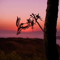 Metalbird Canada's Ruby-Throated Hummingbird is a handcrafted metal silhouette decor, depicting the bird and a flower on a tree trunk, set against a vibrant sunset sky in shades of pink, orange, and purple over a blurred landscape.