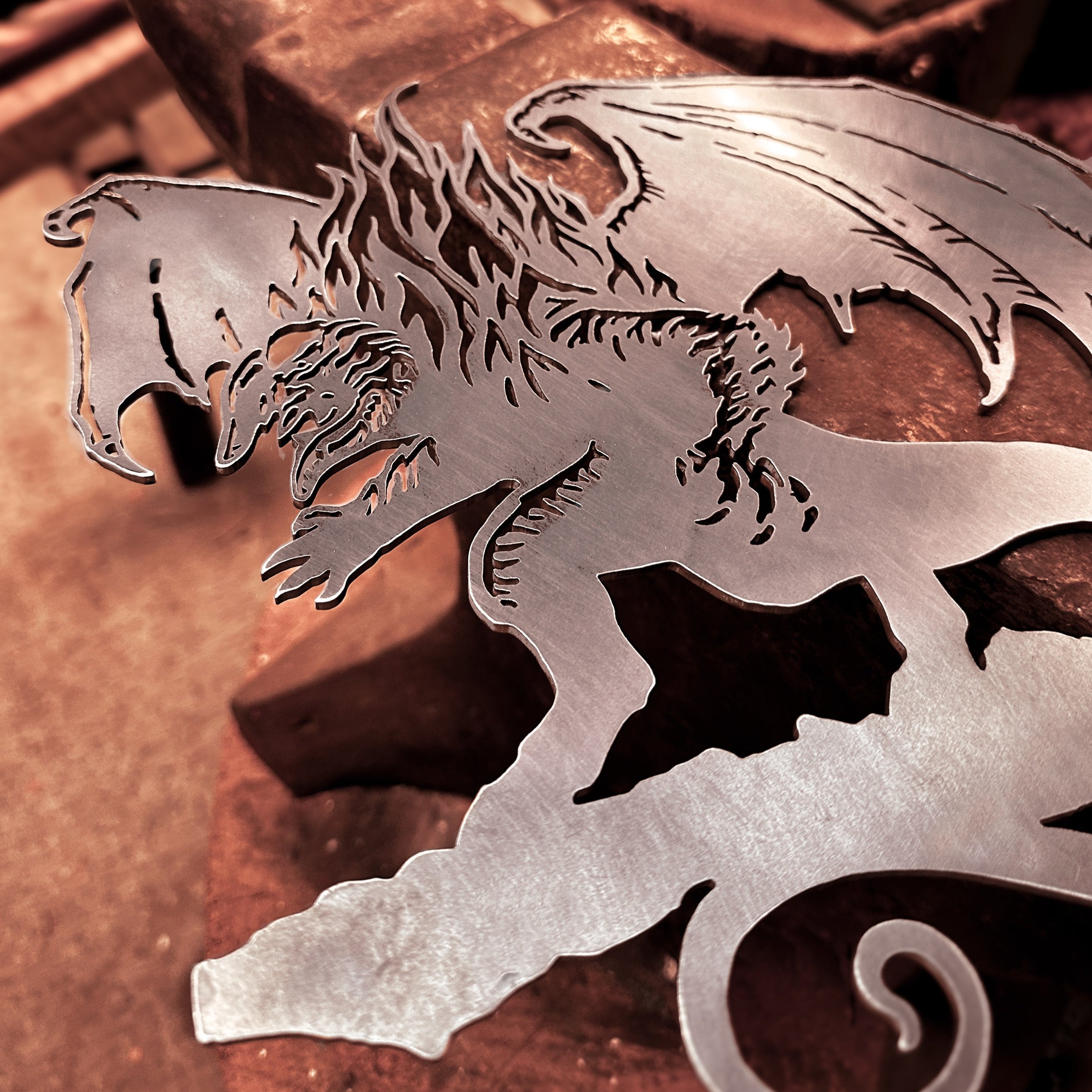 The BALROG™ by Metalbird Canada is a striking corten steel dragon cutout with outstretched wings and claws, perfect for standout garden art. It’s shown on a workbench in a workshop, with blurred tools and anvil in the background.