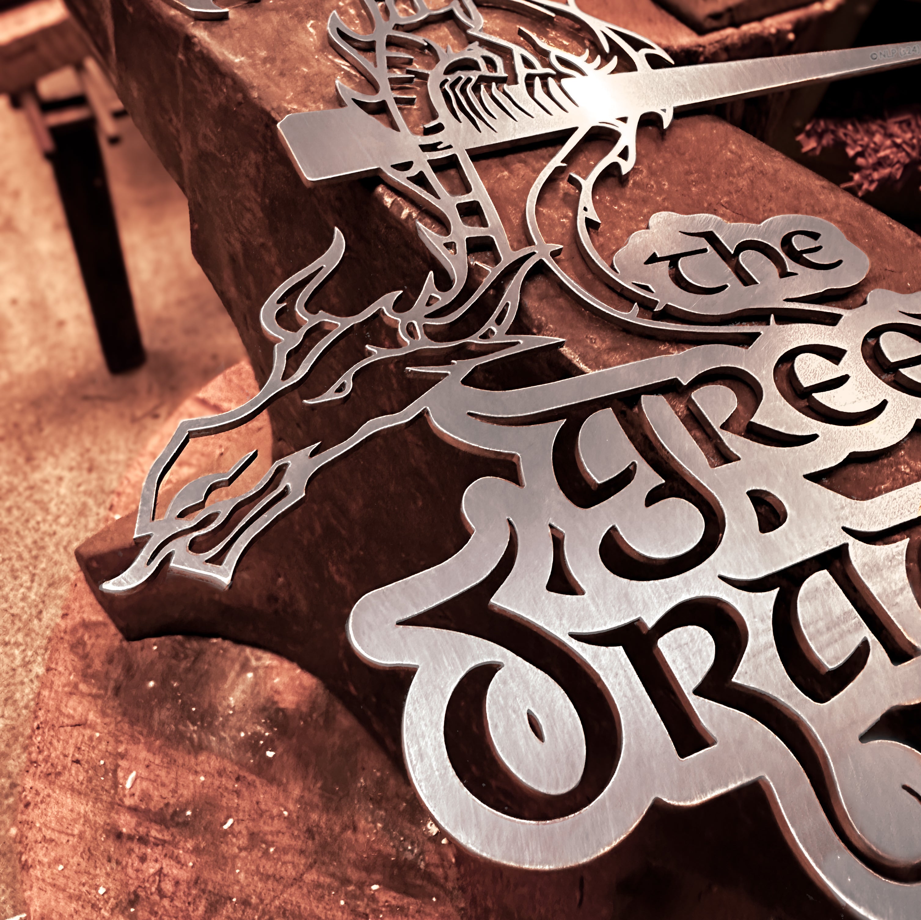 A close-up of THE GREEN DRAGON™, a metallic sign by Metalbird Canada with an artistic dragon and ornate text, displayed on a worn workbench—perfect as unique garden art or a gift for fantasy lovers.