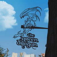 A black corten steel sign by Metalbird Canada, featuring a dragon and the words "THE GREEN DRAGON™," is mounted on a tree against a blue sky with laundry in the background—unique yard art and a great gift.