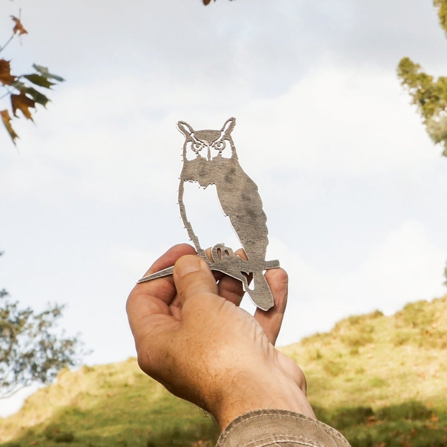 A hand holds up Metalbird Canada’s Mini Owl, a small corten steel cutout shaped like an owl on a branch, with a grassy hillside and cloudy sky in the background.