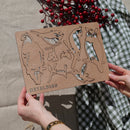 Someone holds a Wooden Hanging Decoration by Metalbird Canada with bird silhouettes like an owl and hummingbird. "METALBIRD" is etched in the wood—unique garden art or a thoughtful gift, pictured over checkered cloth with red berries nearby.