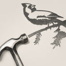 A metal hammer rests beside Metalbird Canada’s “Cardinal,” a finely detailed metal cutout of a bird on a branch—perfect as unique yard art or a striking gift, with intricate features that stand out on any background.