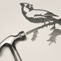 A metal hammer rests beside Metalbird Canada’s “Cardinal,” a finely detailed metal cutout of a bird on a branch—perfect as unique yard art or a striking gift, with intricate features that stand out on any background.
