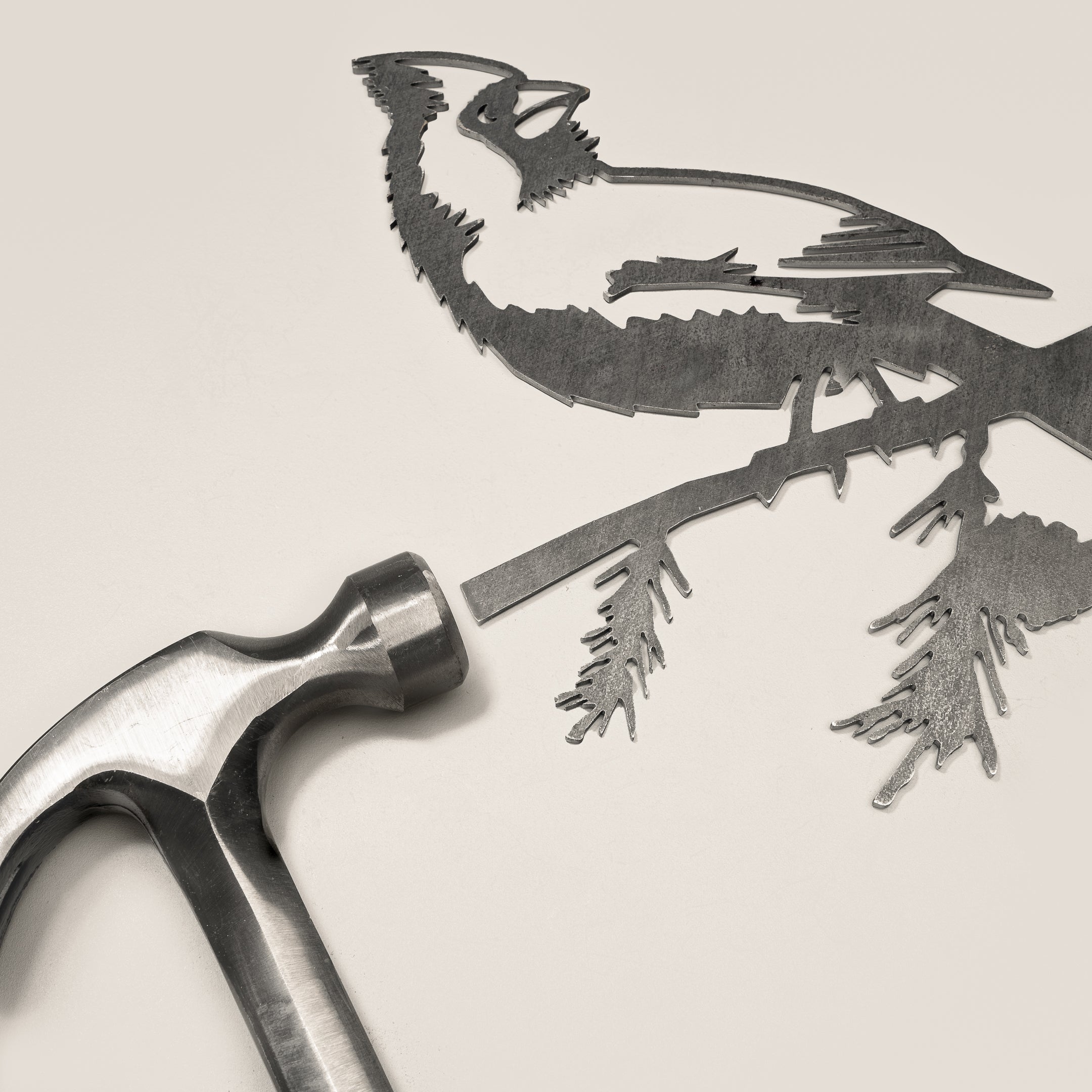 A metal hammer rests beside Metalbird Canada’s “Cardinal,” a finely detailed metal cutout of a bird on a branch—perfect as unique yard art or a striking gift, with intricate features that stand out on any background.