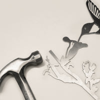 A metal hammer rests beside Metalbird Canada’s "Hummingbird," a corten steel silhouette of a bird, branch, and flower—unique garden art accents—displayed on a light background.