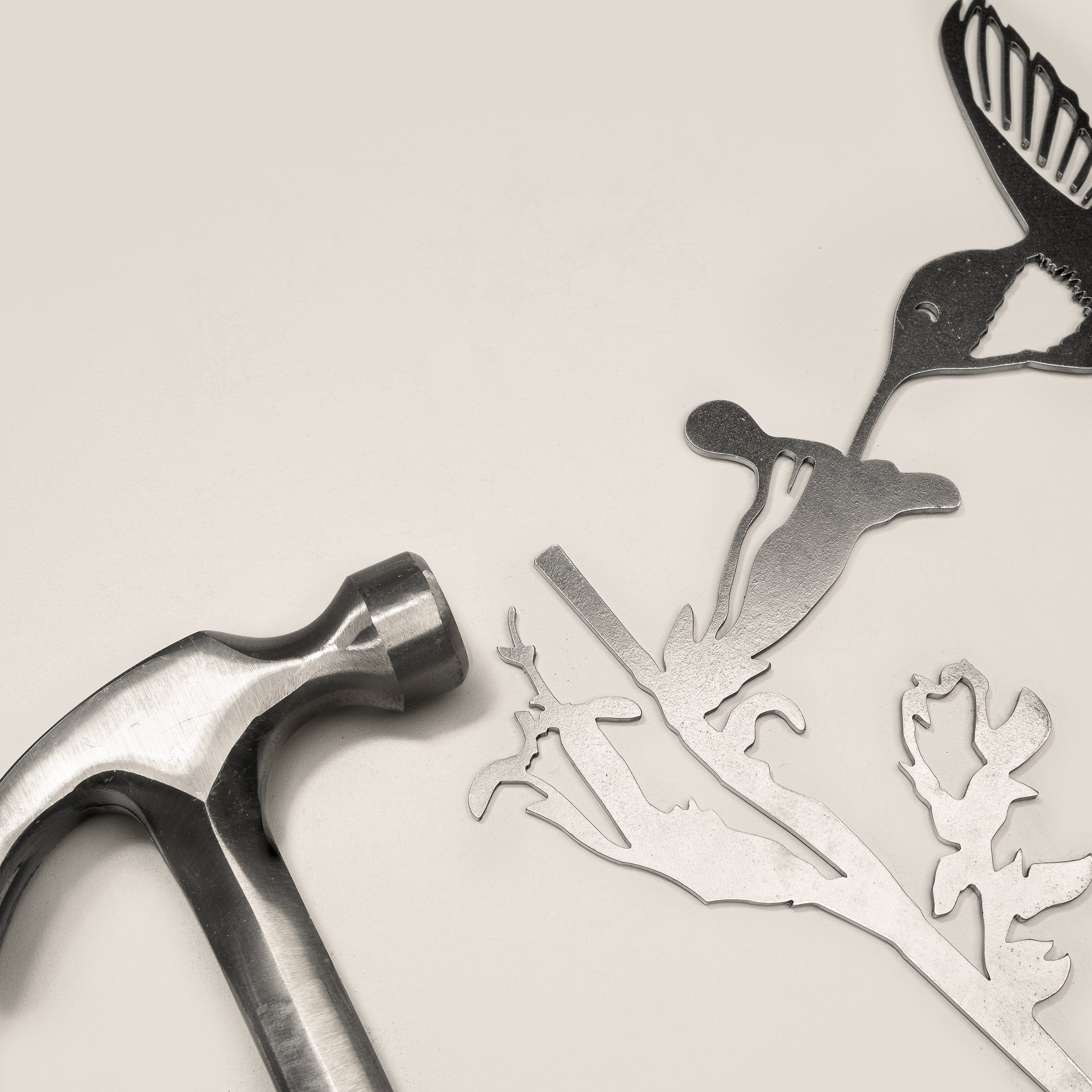 A metal hammer rests beside Metalbird Canada’s "Hummingbird," a corten steel silhouette of a bird, branch, and flower—unique garden art accents—displayed on a light background.