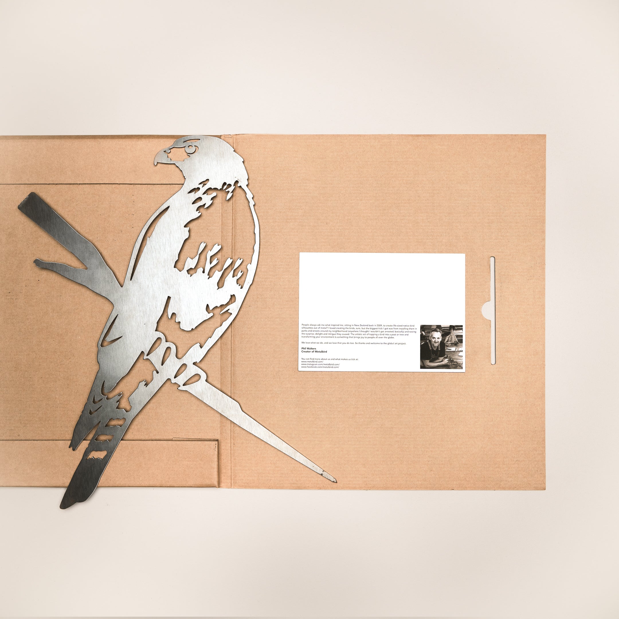 The Metalbird Canada Red-Tailed Hawk metal silhouette rests on an open brown folder with a white card and black-and-white photo inside, making a thoughtful gift or striking yard art accent. Set against a light beige background.
