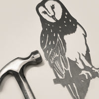 A metal hammer rests beside the Metalbird Canada “Barn Owl,” an intricately cut corten steel silhouette of an owl on a branch, both displayed on a light surface.