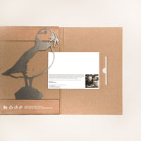The Metalbird Canada Puffin comes in eco-friendly cardboard packaging featuring a puffin silhouette, white icons, and text. An inserted white card with text and a black-and-white photo makes it ideal for unique garden art lovers.