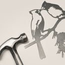 The Metalbird Canada Kissing Cardinals—a delicate metal silhouette of two birds on a branch—rests beside a metal hammer against a neutral background, offering perfect inspiration for garden art enthusiasts.