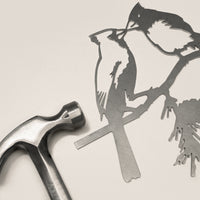 The Metalbird Canada Kissing Cardinals—a delicate metal silhouette of two birds on a branch—rests beside a metal hammer against a neutral background, offering perfect inspiration for garden art enthusiasts.