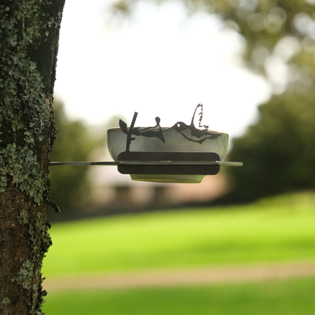 The Bowl Feeder by Metalbird Canada is a small, clear bowl with a corten steel fishing figure on a tree trunk—unique yard art against a grassy field and the perfect gift for any nature lover.