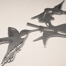 A close-up of Metalbird Canada’s Hummingbird Honeys, a metal garden art piece with two stylized hummingbirds perched on branches, featuring a smooth, reflective surface against a light background.