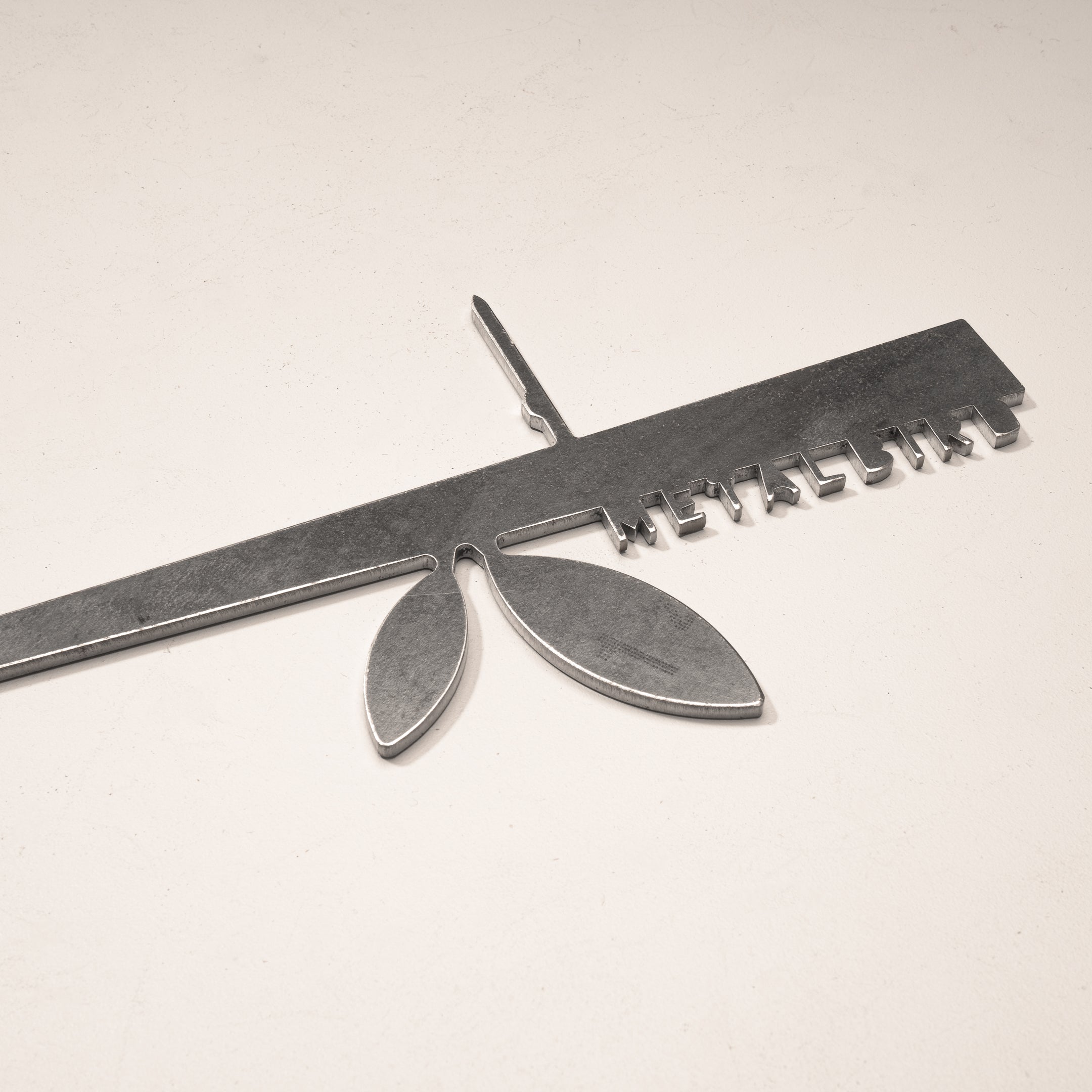 The Stick Bird Feeder by Metalbird Canada features a ruler-shaped metallic design with keys and leaf-like accents, making it unique garden art or a thoughtful gift, shown here on a light-colored surface.
