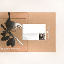 A flat cardboard package contains Metalbird Canada's Wren corten steel bird on a branch art piece, plus a printed card with text and a small black-and-white portrait, all displayed on a light surface.