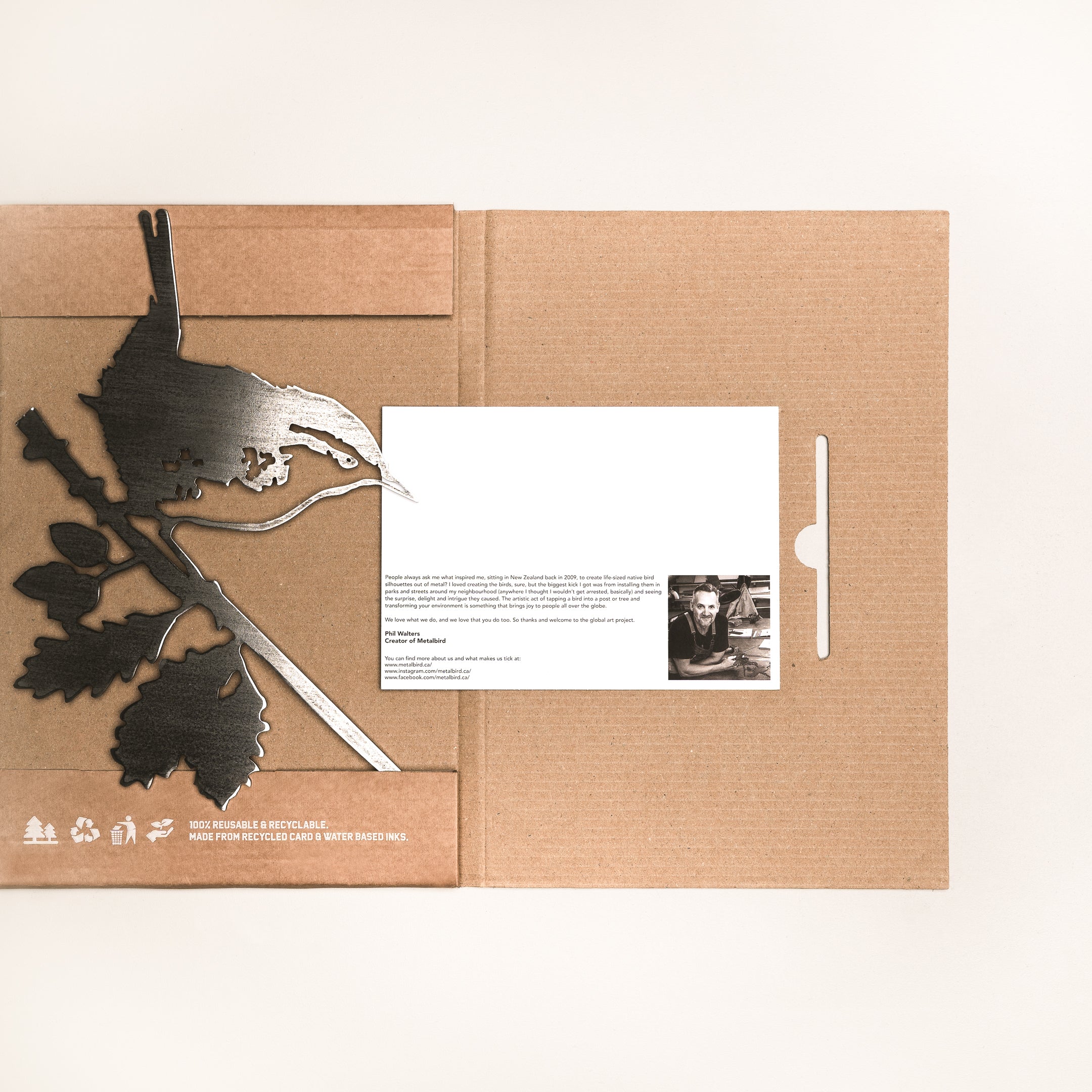 A flat cardboard package contains Metalbird Canada's Wren corten steel bird on a branch art piece, plus a printed card with text and a small black-and-white portrait, all displayed on a light surface.