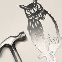 The Great Horned Owl by Metalbird Canada is a detailed metal cutout with lifelike feathers and facial features, shown next to a silver hammer on a light surface—a unique garden art gift for nature lovers.
