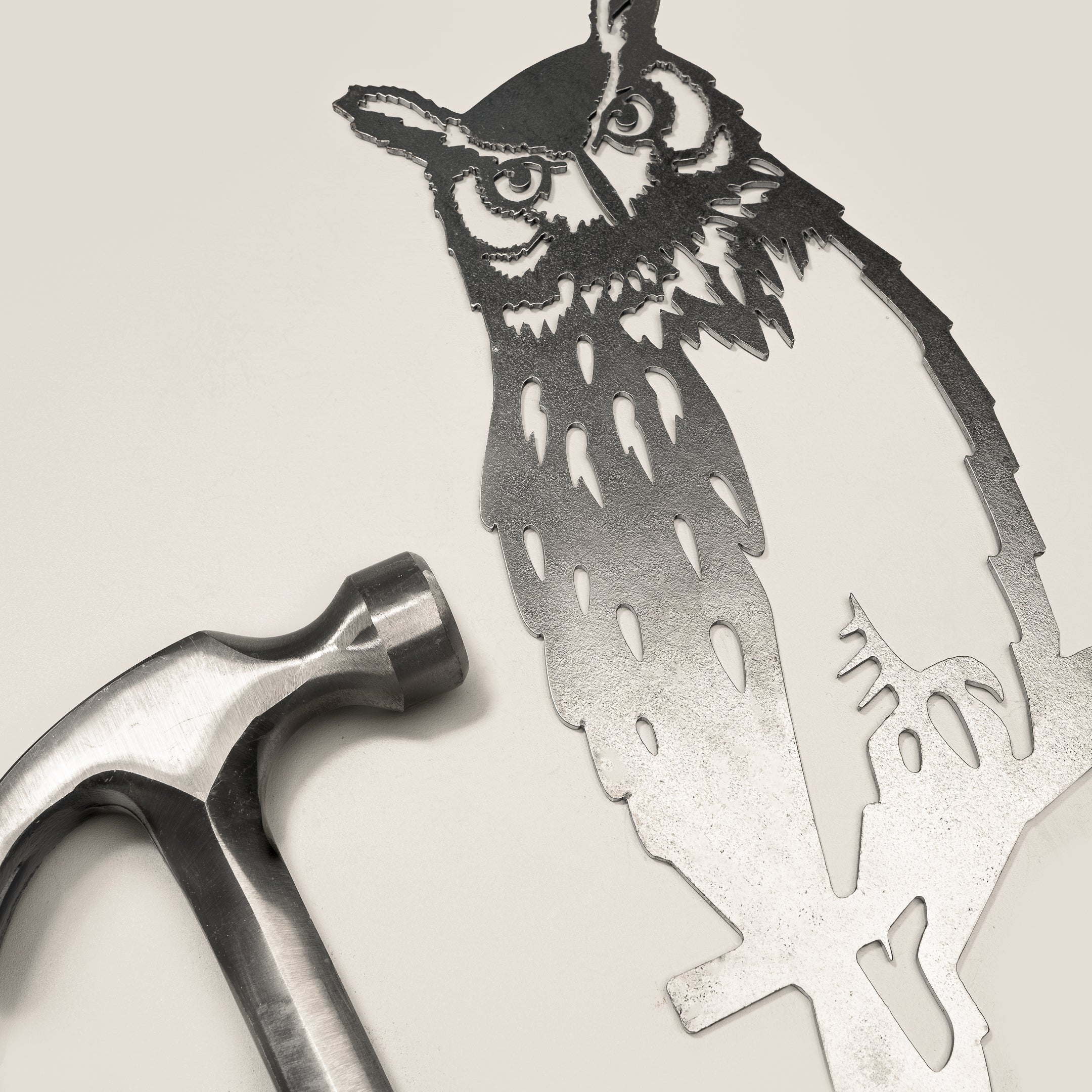 The Great Horned Owl by Metalbird Canada is a detailed metal cutout with lifelike feathers and facial features, shown next to a silver hammer on a light surface—a unique garden art gift for nature lovers.