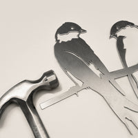 A silver hammer rests beside Metalbird Canada's "Pair of Swallows" metal bird silhouettes perched on a branch, all set against a light background—a thoughtful gift for any occasion.