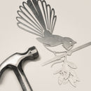 The Fantail / New Zealand Pīwakawaka by Metalbird Canada, a corten steel bird silhouette with floral branch, rests on a light surface beside a silver hammer—an ideal gift for nature lovers.
