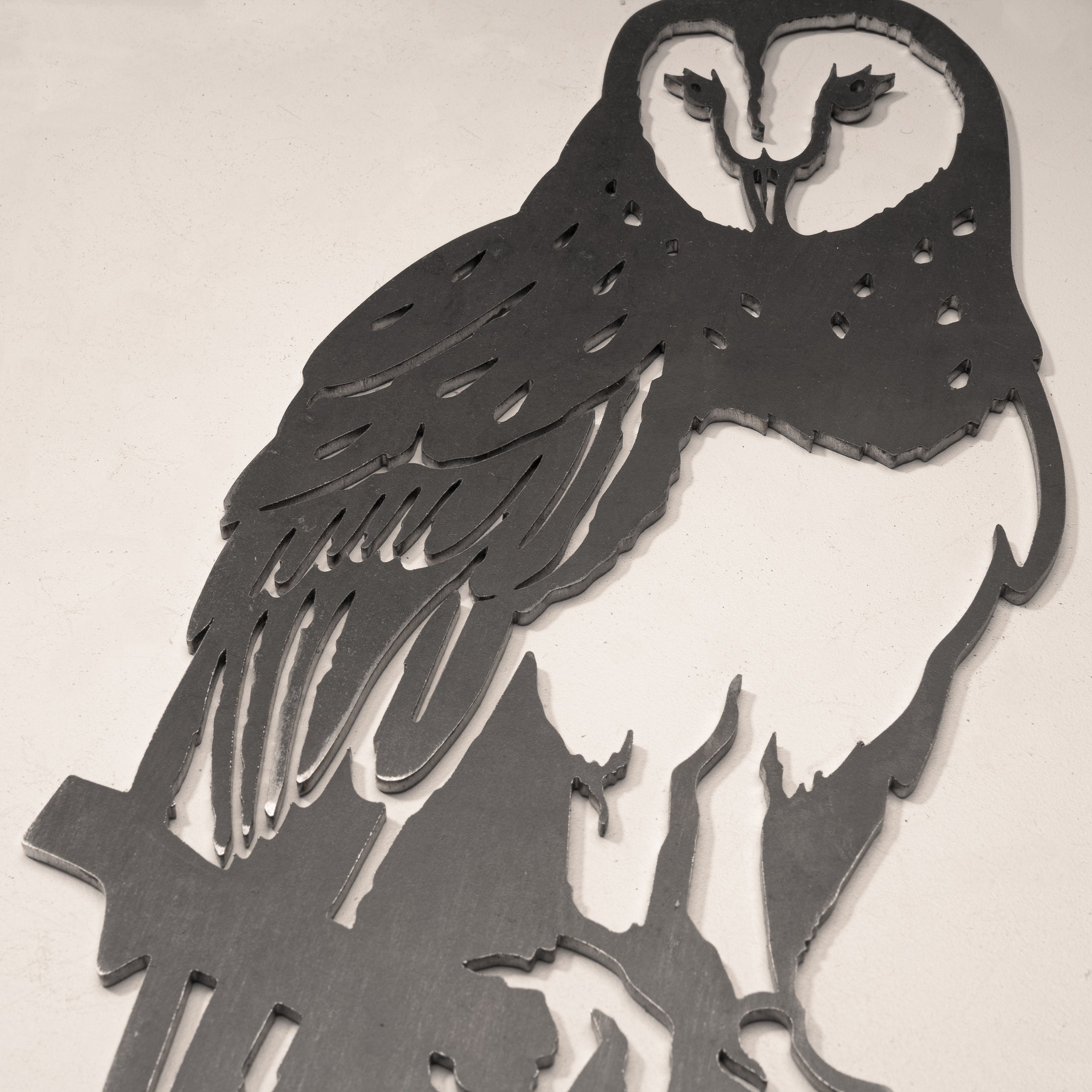 The Barn Owl by Metalbird Canada is a metal wall art piece with a cut-out silhouette of an owl perched on a branch, showcasing feather and facial details against a light-colored wall.