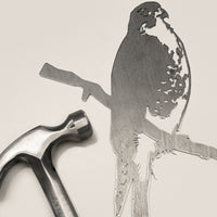 A silver hammer beside Metalbird Canada’s “Peregrine Falcon” corten steel silhouette, both set on a light surface—an ideal gift for nature lovers.