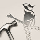 The Metalbird Canada Blue Jay, a stylized metal cutout of a bird on a branch, sits next to a silver hammer on a light background—perfect as unique garden or yard art.