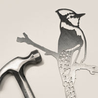 The Metalbird Canada Blue Jay, a stylized metal cutout of a bird on a branch, sits next to a silver hammer on a light background—perfect as unique garden or yard art.