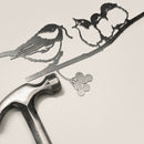 Chickadee & Chicks" by Metalbird Canada—a striking metal art piece crafted from thin corten steel—features three birds on a branch, shown with a hammer nearby; ideal as unique garden decor or a thoughtful gift.