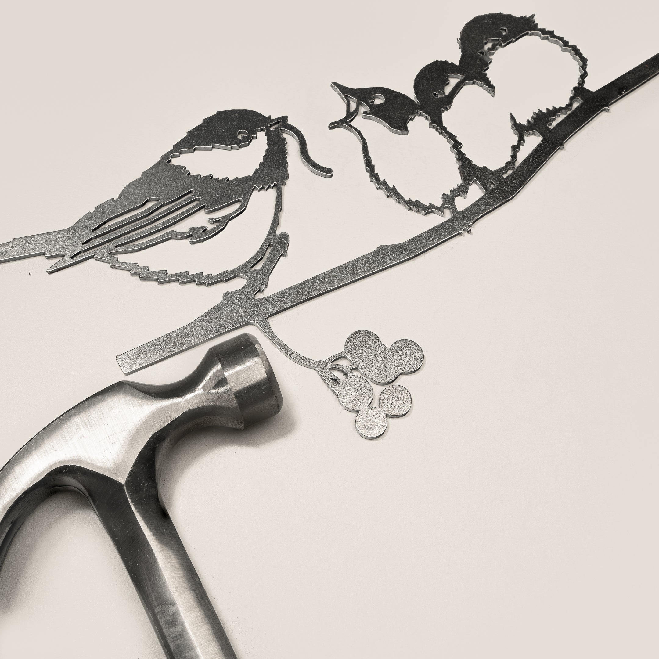 Chickadee & Chicks" by Metalbird Canada—a striking metal art piece crafted from thin corten steel—features three birds on a branch, shown with a hammer nearby; ideal as unique garden decor or a thoughtful gift.