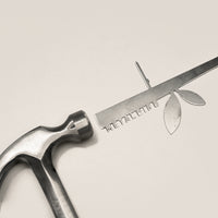 A Metalbird Canada Stick Bird Feeder, shaped like a twig with leaves and text cutouts, is shown next to a metal hammer on a plain light background. Both resemble garden or yard art.