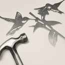 The Hummingbird Honeys by Metalbird Canada features two corten steel hummingbirds and a flower beside a metal hammer, highlighting the contrast between delicate art and sturdy tool—perfect for nature lovers.