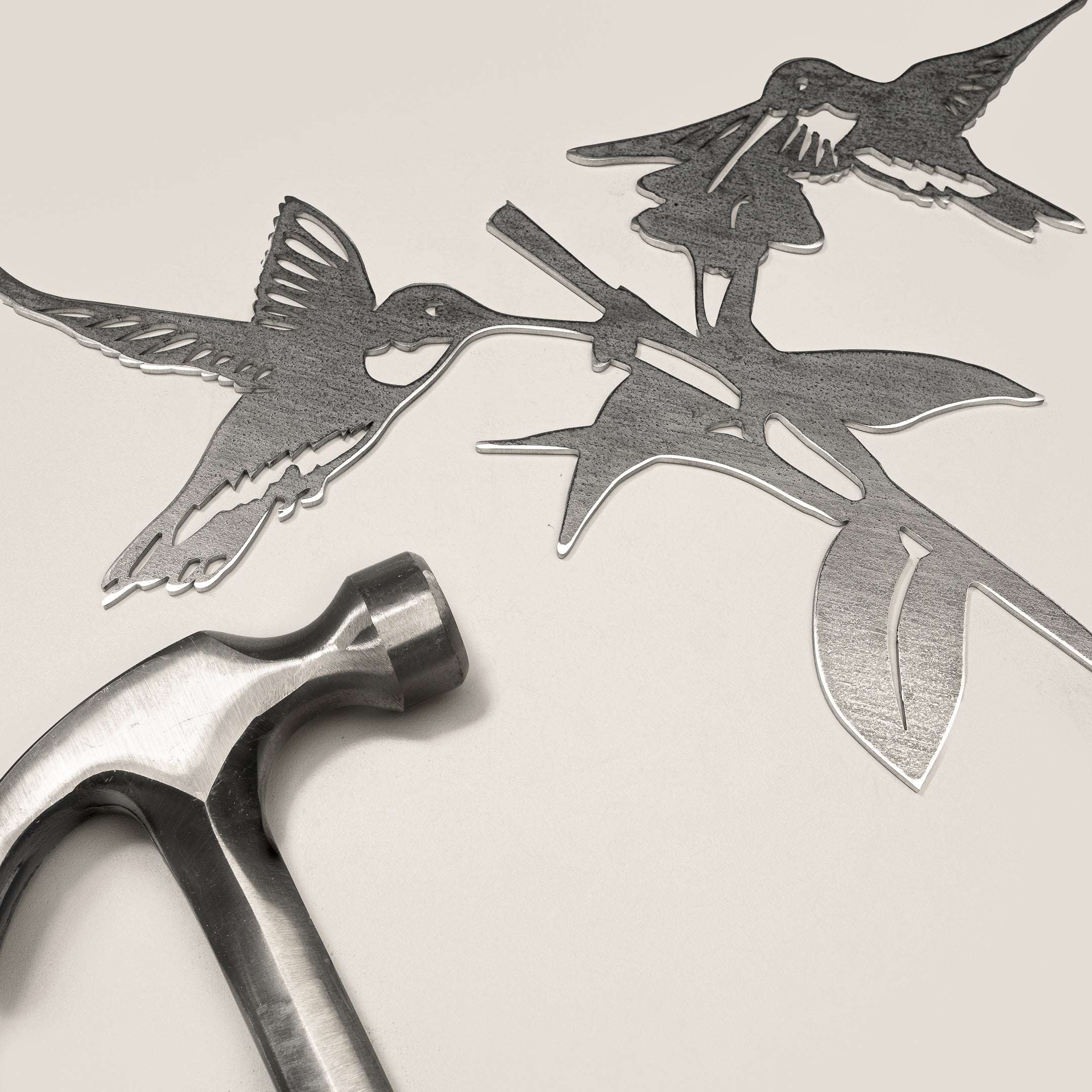 The Hummingbird Honeys by Metalbird Canada features two corten steel hummingbirds and a flower beside a metal hammer, highlighting the contrast between delicate art and sturdy tool—perfect for nature lovers.