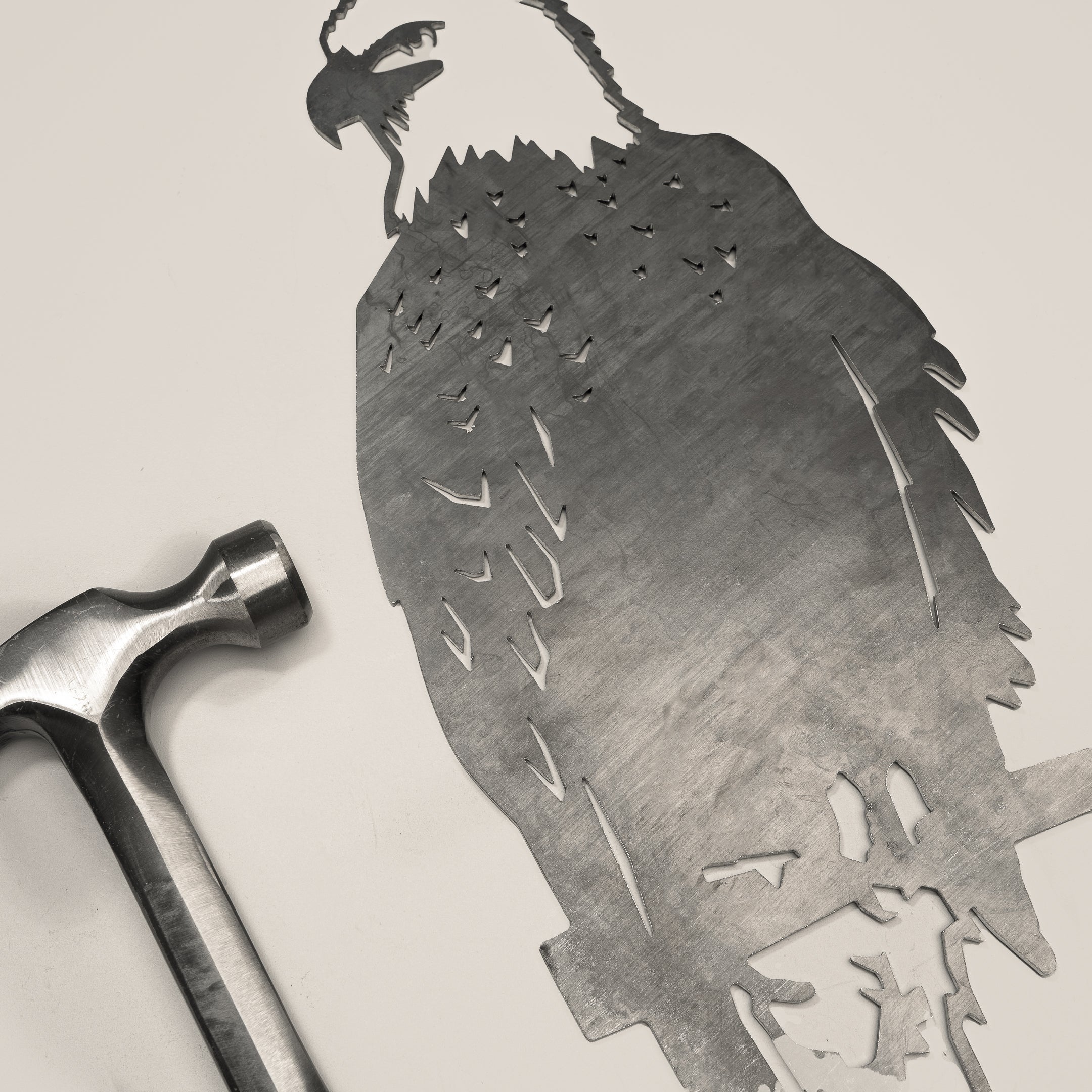 A metal hammer rests beside Metalbird Canada's Bald Eagle—a flat, intricately cut corten steel piece shaped like an eagle on a branch—ideal as unique garden art or a thoughtful gift, both set on a light surface.