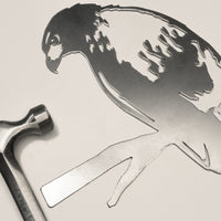 A Red-Tailed Hawk metal silhouette by Metalbird Canada lies next to a metal hammer on a light surface. This detailed yard art makes a unique gift and adds character to any outdoor space. The hammer is positioned left of the artwork.